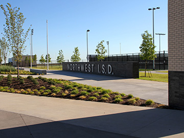 A low brick wall with a sign for Northwest ISD in Texas, positioned outside of a school building and athletic fields